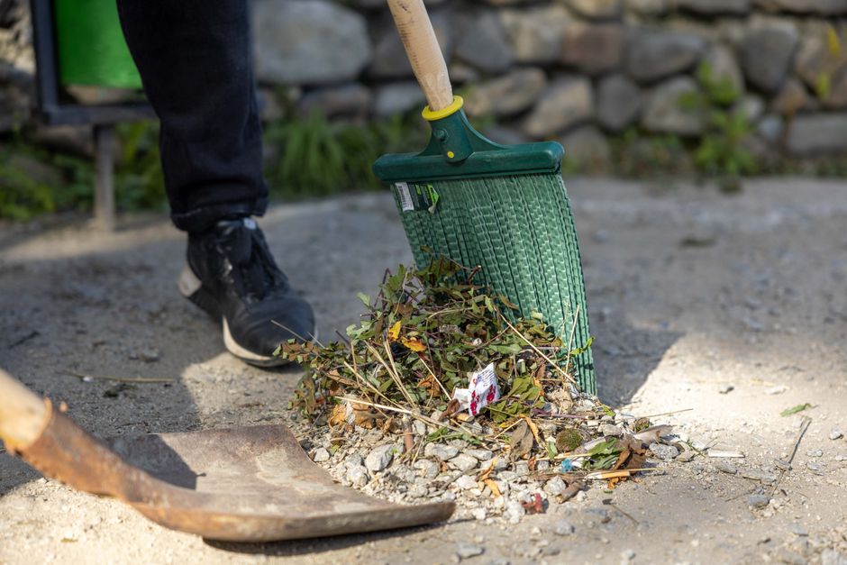 A person wearing black trousers and black sneakers is using a green plastic garden rake with a wooden handle to gather fallen leaves, small twigs, and garden debris into a pile on a compacted gravel s