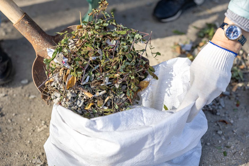 A person wearing black trousers and black sneakers is using a green plastic garden rake with a wooden handle to gather fallen leaves, small twigs, and garden debris into a pile on a compacted gravel s