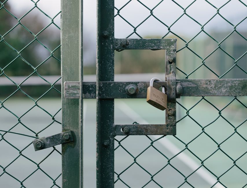A close-up of a green metal garden gate with a lock securing the latch, set against a backdrop of a tennis court surface. The gate features a chain-link design, with visible rust and weathering on the